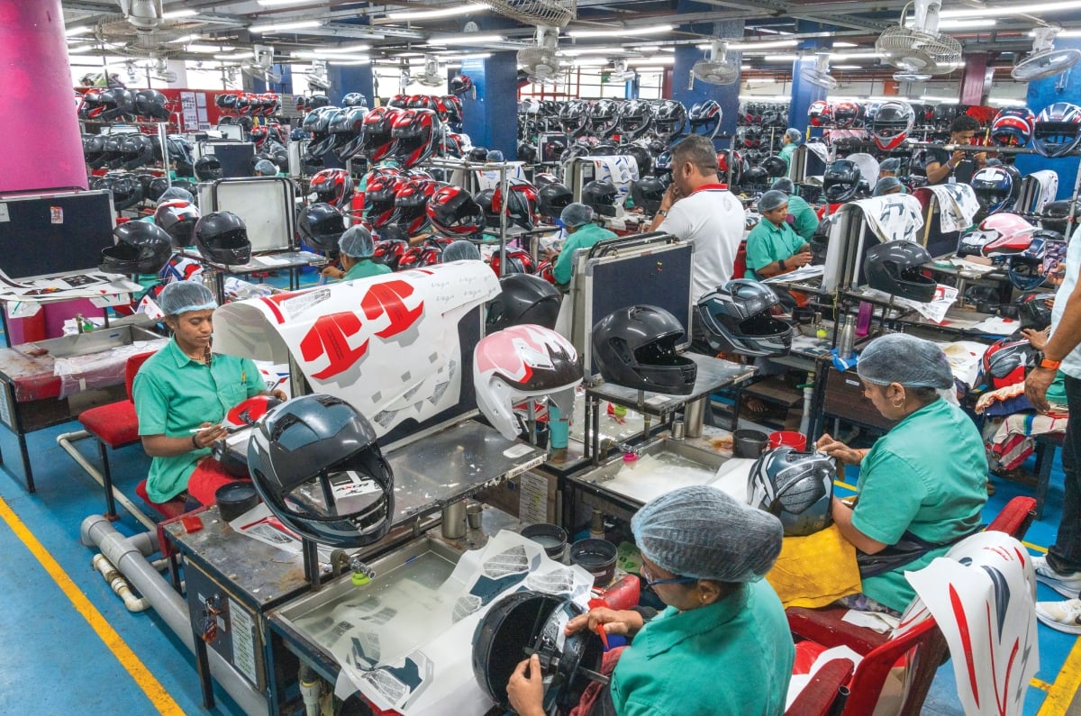 Vega helmet manufacturing facility with workers assembling and finishing motorcycle helmets at multiple workstations, surrounded by rows of helmets and production equipment.